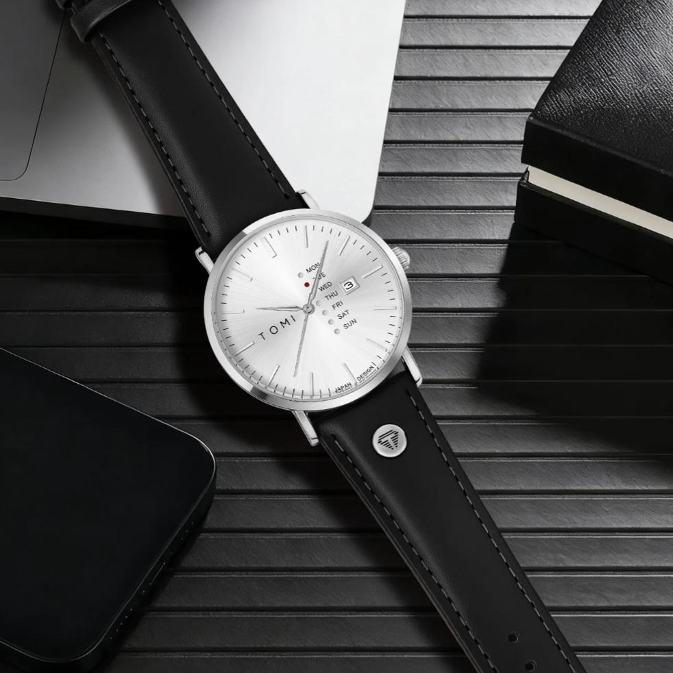 Montre TOMI à cadran blanc et bracelet en cuir noir posée à plat à côté d'un téléphone et d'un carnet noirs