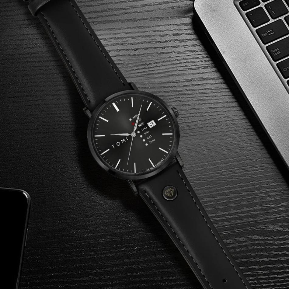 Montre TOMI noire à cadran noir et bracelet en cuir noir posée sur un bureau en bois sombre près d'un ordinateur portable